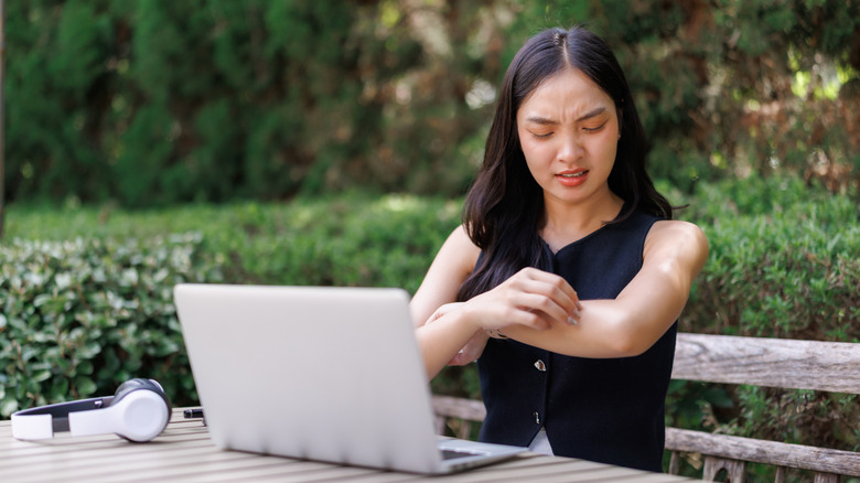 Young woman working outside on laptop itching mosquito bite