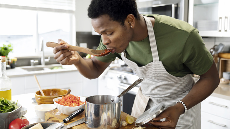 A person cooking in a kitchen, tasting food from a silver pot