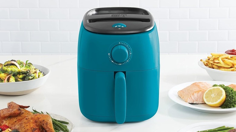 A teal blue DASH Tasti-Crisp Compact Air Fryer sits on a white countertop surrounded by platters of food