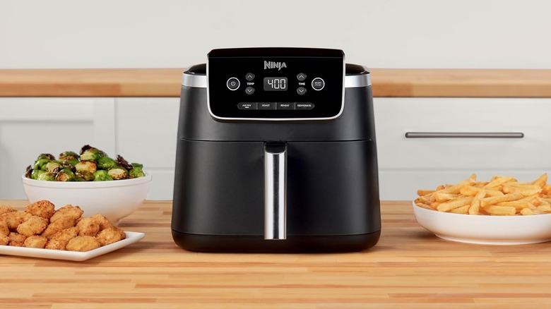 A Ninja air fryer is surrounded by platters of chicken fingers, Brussels sprouts, and french fries