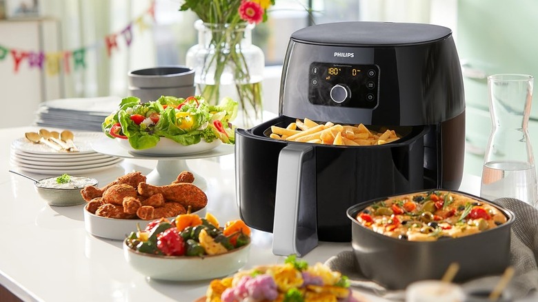 A Phillips Air Fryer basket is half opened to reveal cooked french fries while sitting on a table surrounded by bowls of party food