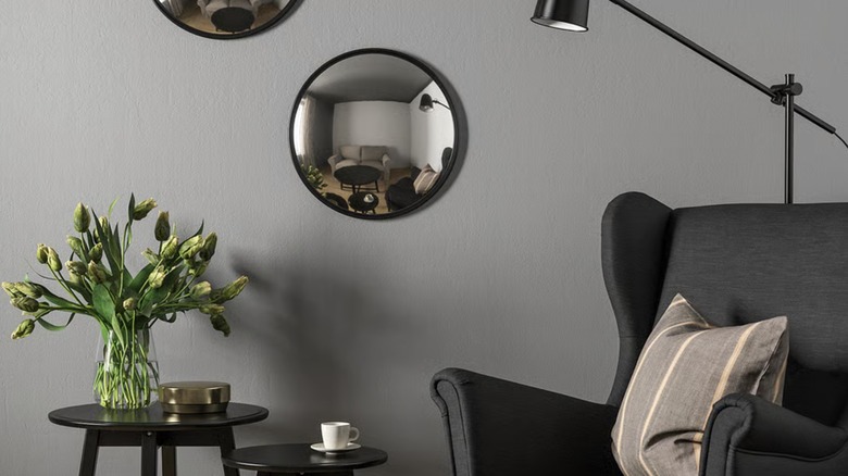 A convex mirror hangs on a wall above a black armchair and vase full of flowers