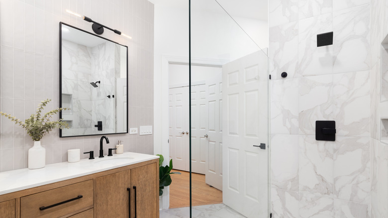 A white marble bathroom with wooden cabinets, black accents on mirror and hardware, and two green plants