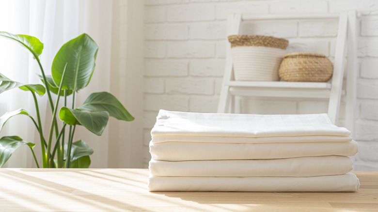 A clean, folded sheet set on a wooden tabletop in a room with a houseplant and a white ladder shelf.