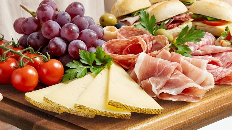 The Kikcoin Acacia Wood Cutting Board being used as a charcuterie board with cheeses, meats, sandwiches, olives, grapes, and tomatoes on top