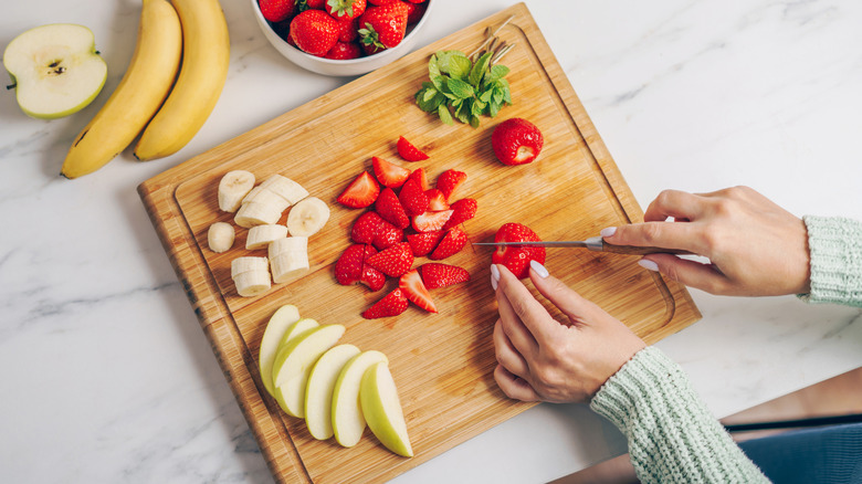 A person cutting up a banana, strawberries, and apple on a wooden cutting board