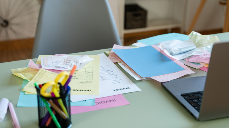 A light green office desk with colored papers, labeled invoice, and a laptop computer.