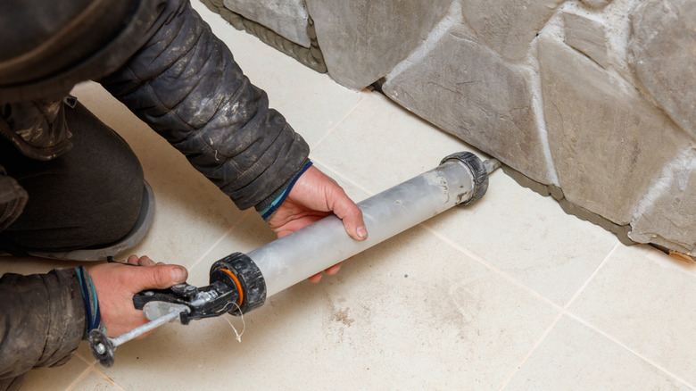 A caulk gun is used to apply gray sealant in the cracks between a tiled floor and cobbled rock wall.