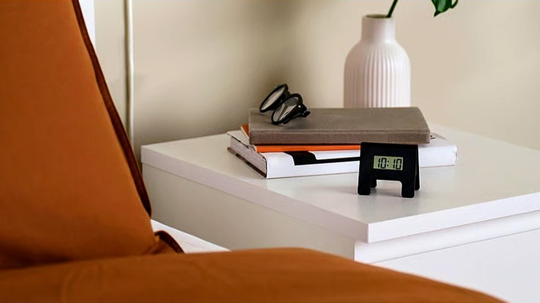 A small black alarm clock sitting on a white bedside table next to a stack of books and a pair of glasses