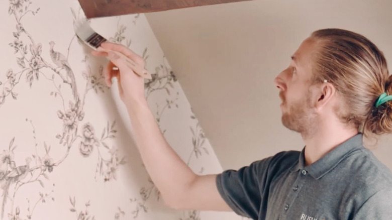 Man uses paintbrush to apply a topcoat to the edges of wallpaper.