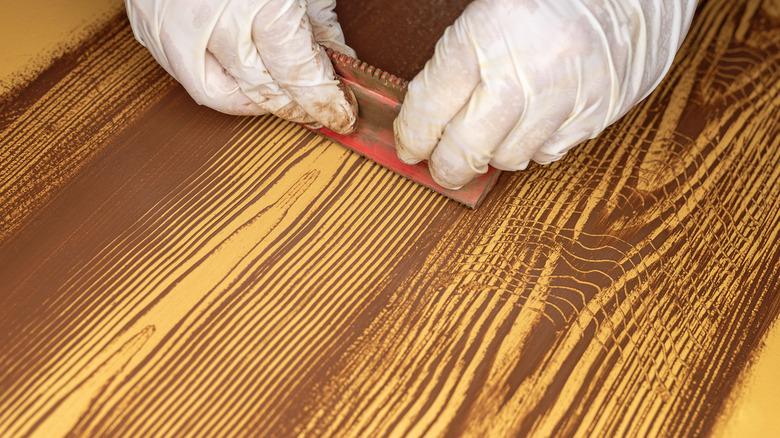 Someone wearing white latex gloves, using a red tool to apply wax to a surface for faux wood grain