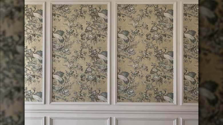 A cream colored wall with wainscotting and picture frame molding with greenish wallpaper featuring birds, flowers, and vines.