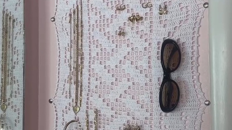 A crochet square tacked to bathroom wall next to mirror holds accessories such as earrings, necklaces and a pair of sunglasses