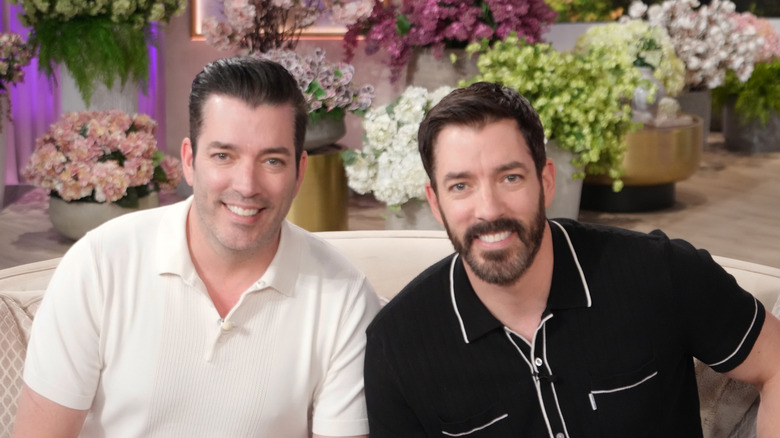 Property Brothers Drew and Jonathan Scott sitting on a loveseat in front of potted flowers.
