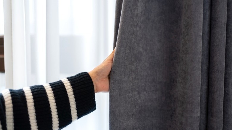 person in black and white sweater pulling gray blackout curtain over sheer curtain
