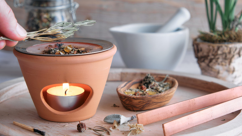 A hand placing a sprig of dried rosemary on top of an incese burner with other containers on a wood surface.