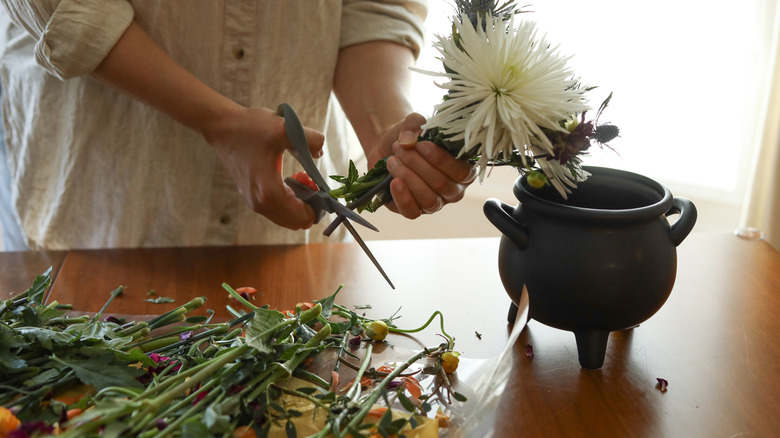 A woman working at a table next to a cauldron-style vase trims the end of the stems of a bunch of cut flowers with scissors.