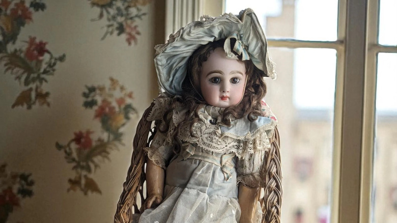 A Jumeau doll sitting on a small cane chair in a room with floral wallpaper and French doors.