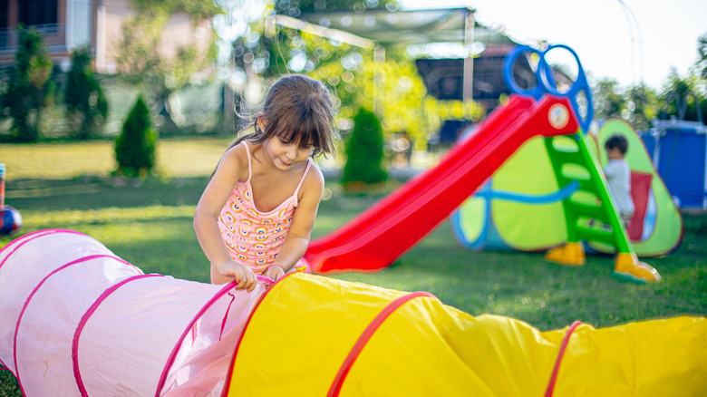Little girl folding up a pop up play tunnel with plastic slide and boy playing in pop up tents behind her