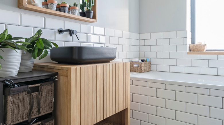 A bathroom with a black sink on top of a natural wood cabinet with a white tub and white tiles in the background