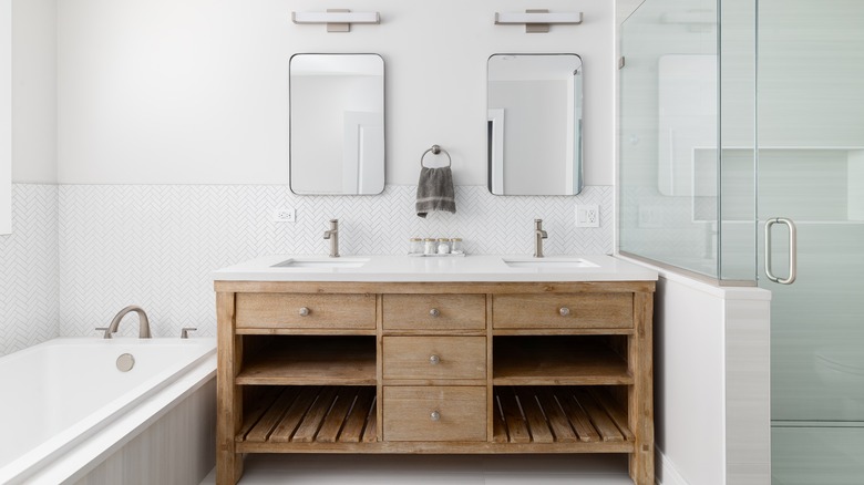 A bathroom with a natural wood cabinet with a white marble countertop, white herringbone tile around the tub, and a walk-in shower with large tiles.