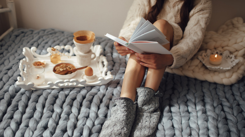 Young woman reading on her bed with a knitted throw, a candle, and tray.