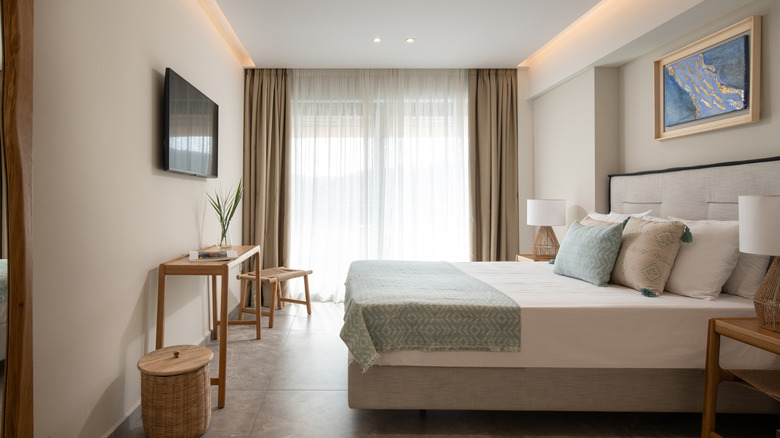 A serene beige-and-white bedroom featuring a cozy bed, a piece of art, a wall-mounted LCD, and a wooden table positioned beneath the screen