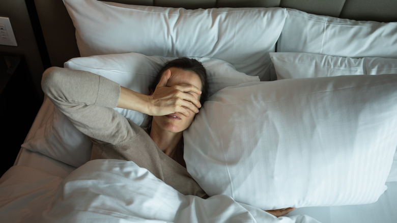 A woman in a gray shirt lying in bed with her hand over her face looking distressed