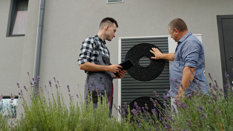An HVAC technician and a homeowner stand outside while looking at a heat pump