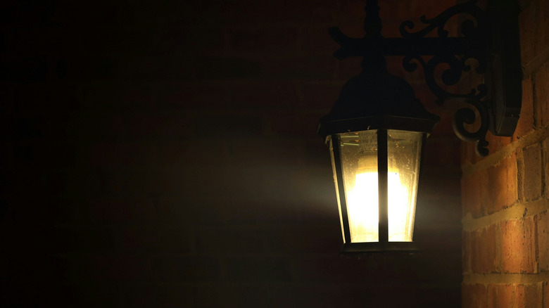 A single outdoor wall-mounted lantern glows against a brick wall at night, casting light into the surrounding dark space.