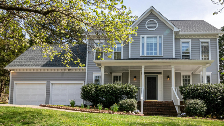 A two-story gray house with white trim and a large front porch, surrounded by trees and shrubs, with a neatly maintained lawn in the foreground.