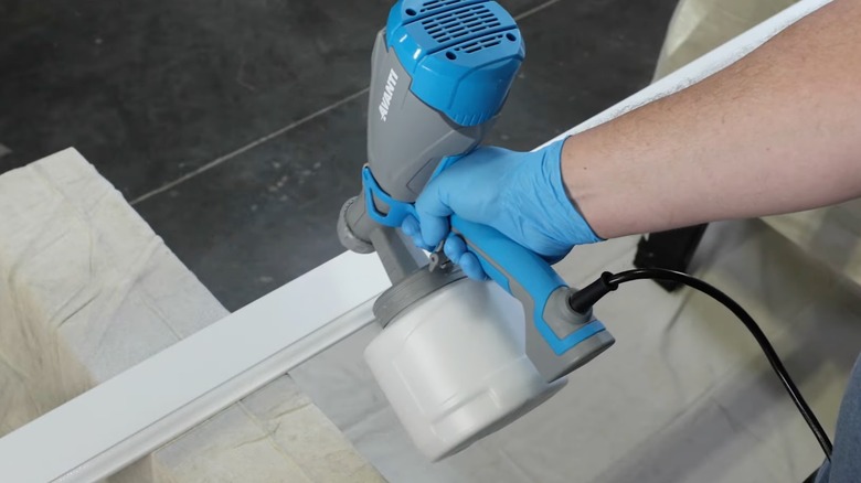 Gloved hand using the Avanti handheld paint sprayer to paint a wooden lawn chair over a drop cloth near pink flower bushes