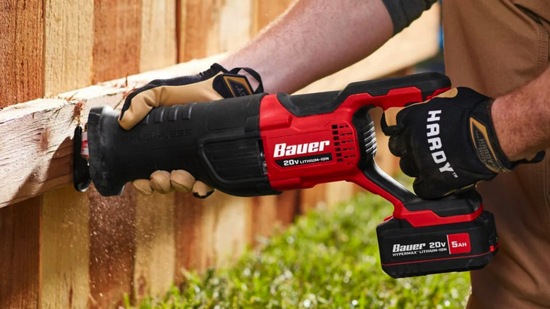 Gloved hands using a Bauer Reciprocating Saw with a 20v battery on a wooden fence over mowed grass