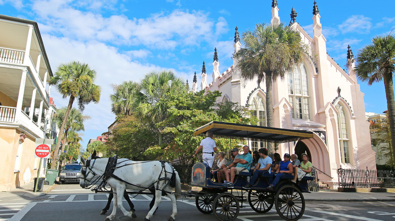 Carriage full of passengers drawn by horses in front of white church and palm trees.