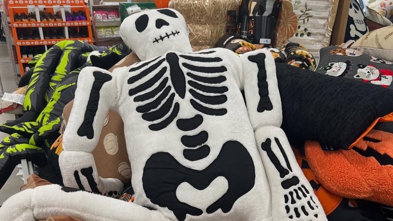 A life-sized skeleton plushie on a stack of other pillows and Halloween decor in a store