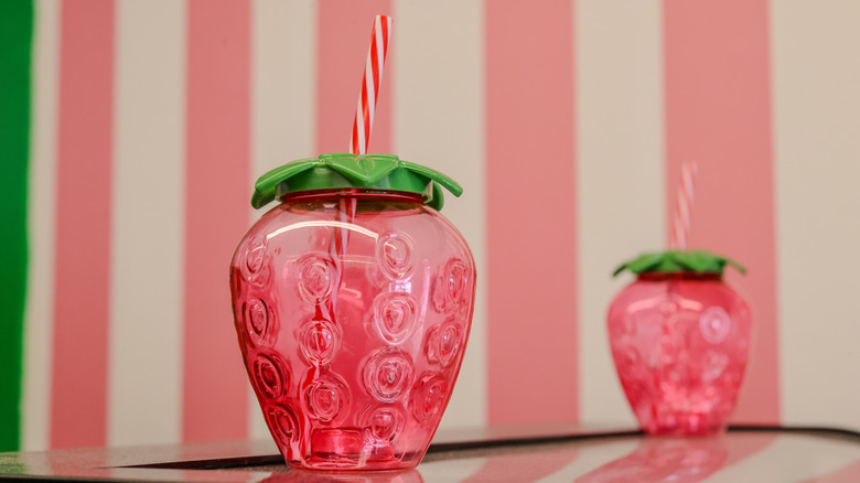 Two strawberry cups with lids and striped straws on a table in front of a pink and white striped wall