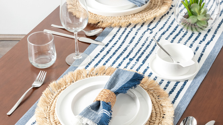 A brown dining table with a blue and white table runner, woven placemats, and blue and white napkins