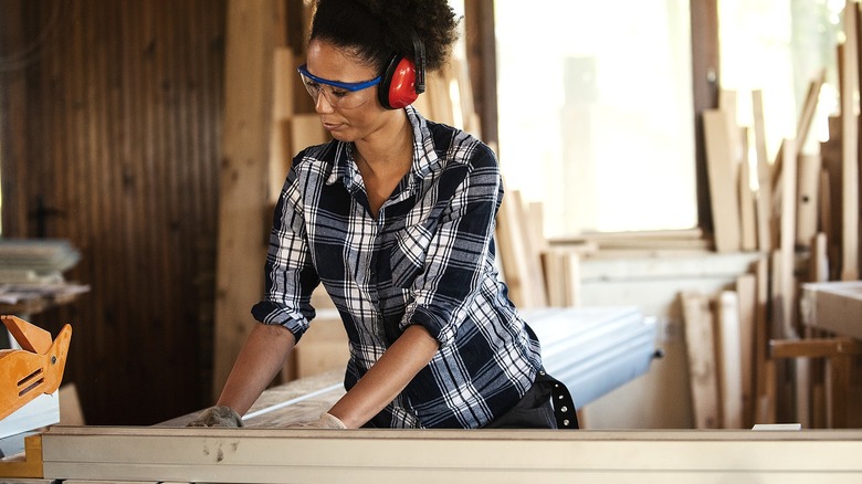A young Black woman wearing eye and ear protection cuts wood in a workshop.