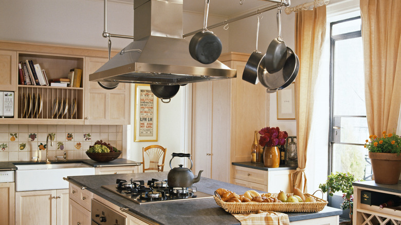 Rustic-looking simple kitchen with light wood cabinets, dark countertop on the island, and soft light brown curtains on the window.