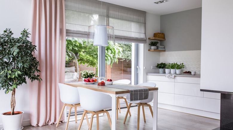 Gray Roman shades and a pink curtain on big, glass windows in a modern kitchen and dining room interior with a wooden table and white chairs