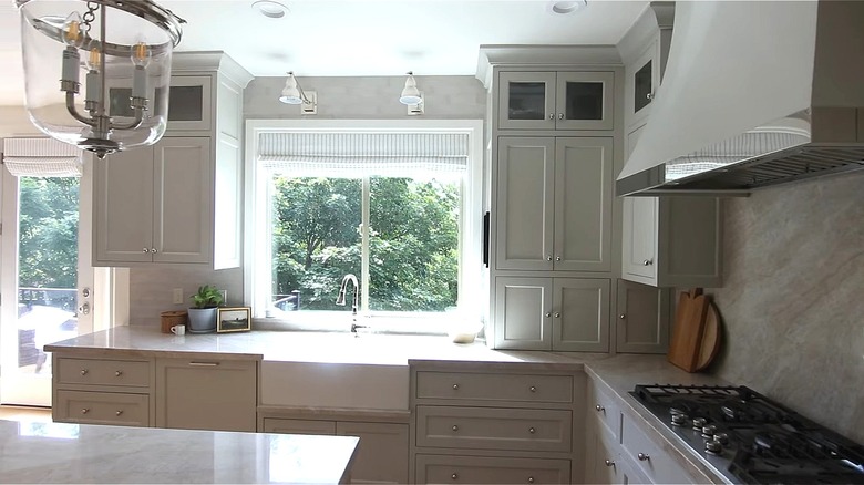 Kitchen space with white cabinets, large window behind sink, with Roman shades.