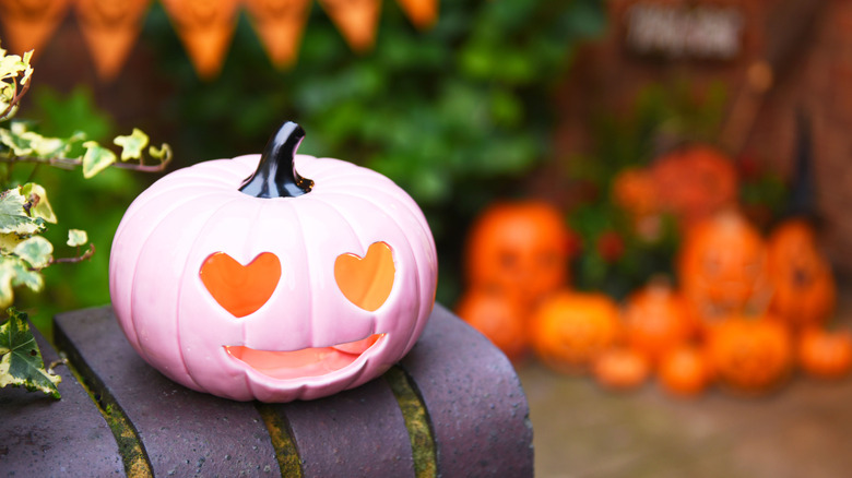A ceramic pink Jack o'lantern wth heart-shaped eyes sitting outside with orange flags and pumpkins in te background