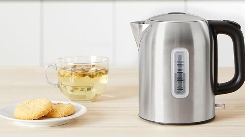 A stainless steel electric kettle beside a cup of tea and plate of biscuits