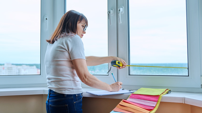 A woman wearing glasses is measuring a window and writing down the results on a notepad with colorful swatches nearby.