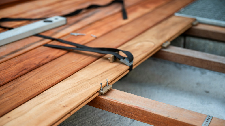 A close-up of wooden decking being installed with hidden fasteners and tools. Wooden planks are securely attached to joists with screws, straps, and brackets.