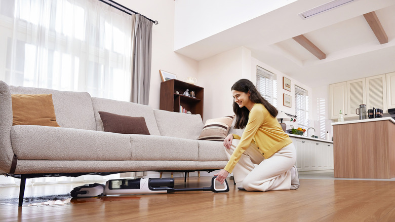 A woman smiles while using a Tineco Floor One Switch S6 to clean under a boucle sofa