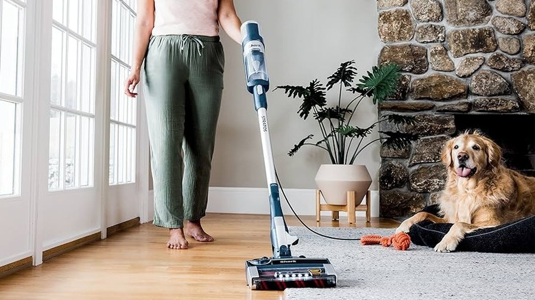 A woman vacuuming with the Shark Ultralight half on a rug and half on a wood floor. There is a golden retriever lying in a bed behind the vacuum.