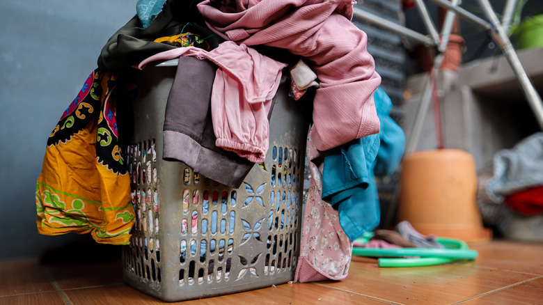 An overflowing pile of laundry sits in a gray basket on a crowded wood floor
