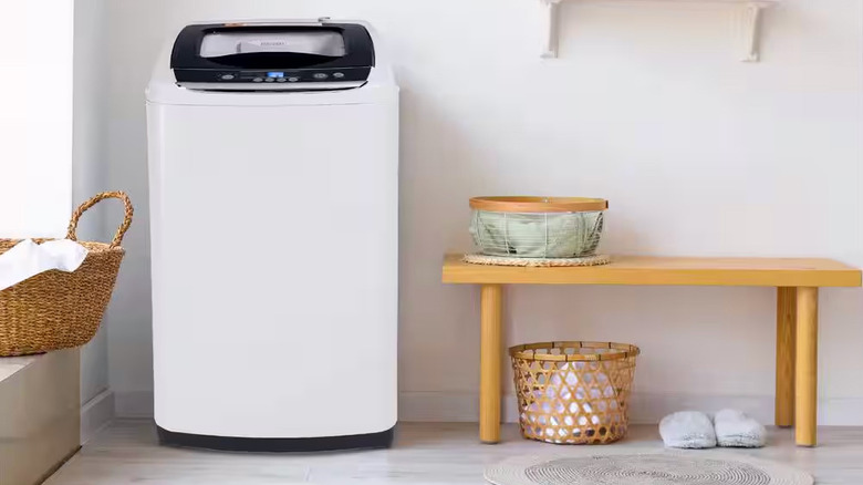 A small top-loading washing machine sits between a wicker laundry basket and small wooden bench