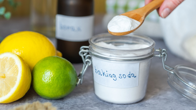Hand holding a scoop of baking soda, with lemons and vinegar in the background to be mixed.